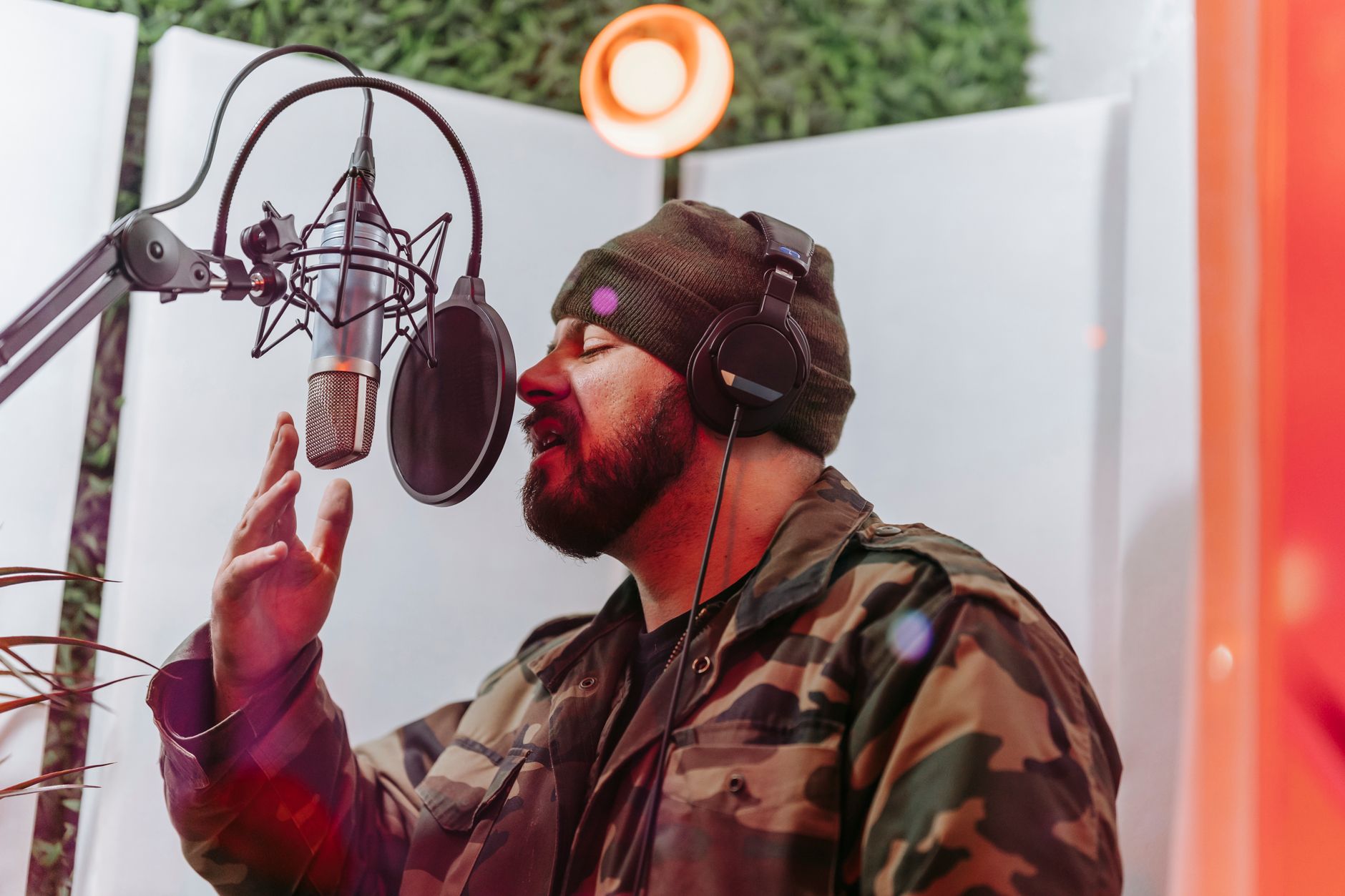Male musician in recording studio singing into a microphone, wearing headphones and camouflage jacket.