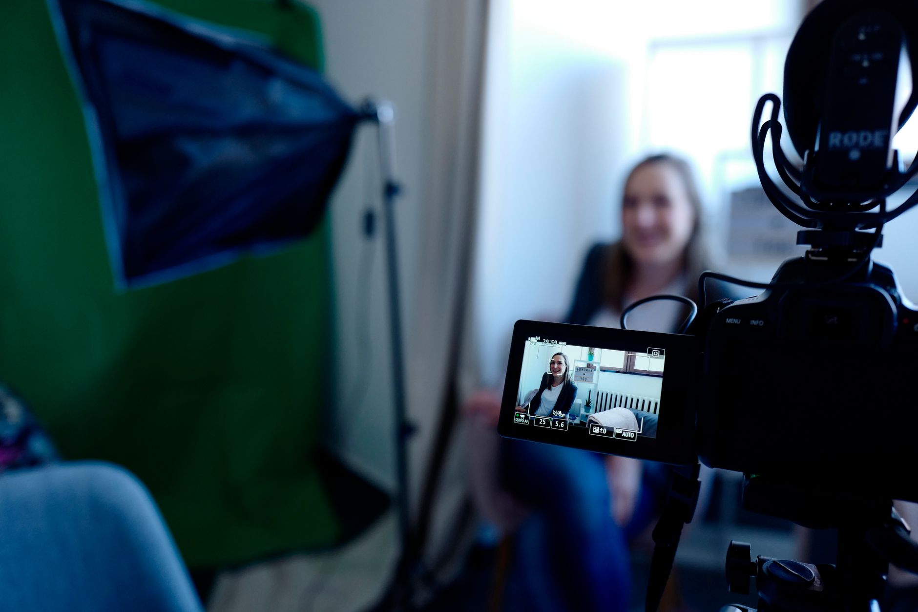 Caméra sur trépied avec micro et écran de retour filmant une femme assise, à côté d’un projecteur softbox et d’un fond vert.